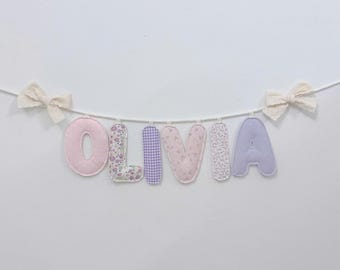 May include: A decorative banner spelling the name OLIVIA with individual fabric letters suspended on a white cord. The letters feature varied patterns including solid pink, floral prints, purple gingham, and light purple with white polka dots.