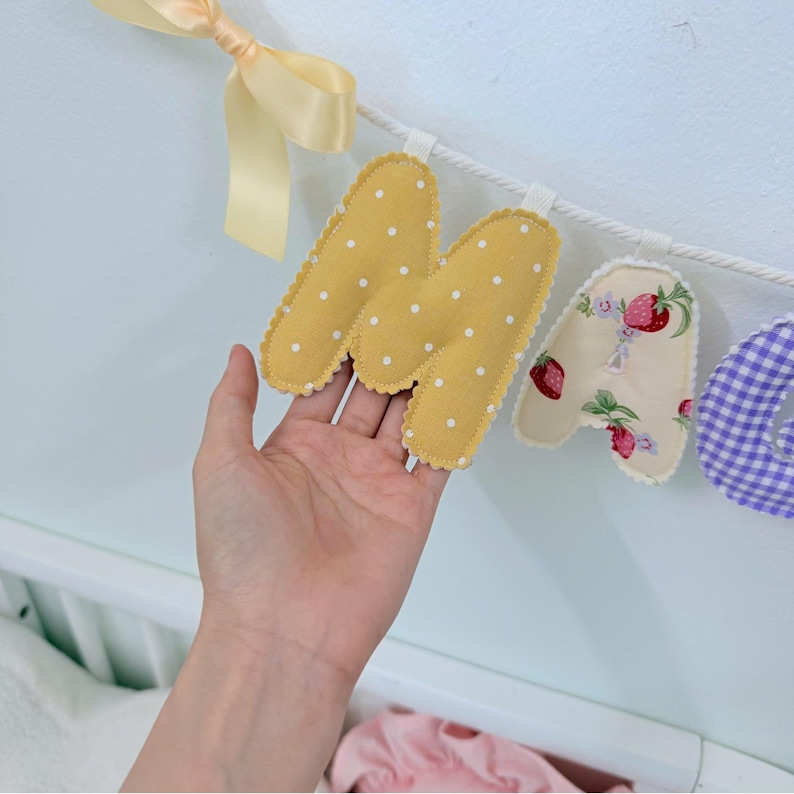 May include: A fabric letter banner featuring a mustard yellow letter M with white polka dots, a cream letter A with a strawberry print, and a purple gingham letter, all suspended from a white rope with a pale yellow ribbon bow.