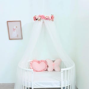 Puede incluir: Una cuna blanca con un dosel blanco transparente colgado de un marco circular adornado con flores artificiales rosas y crema. Dentro de la cuna hay dos cojines decorativos: uno rosa claro en forma de coraz&oacute;n con volantes y otro rosa claro en forma de mariposa.