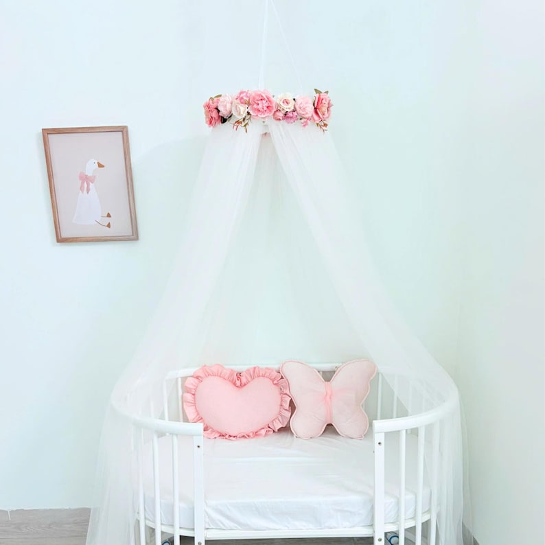 Puede incluir: Una cuna blanca con un dosel blanco transparente colgado de un marco circular adornado con flores artificiales rosas y crema. Dentro de la cuna hay dos cojines decorativos: uno rosa claro en forma de coraz&oacute;n con volantes y otro rosa claro en forma de mariposa.