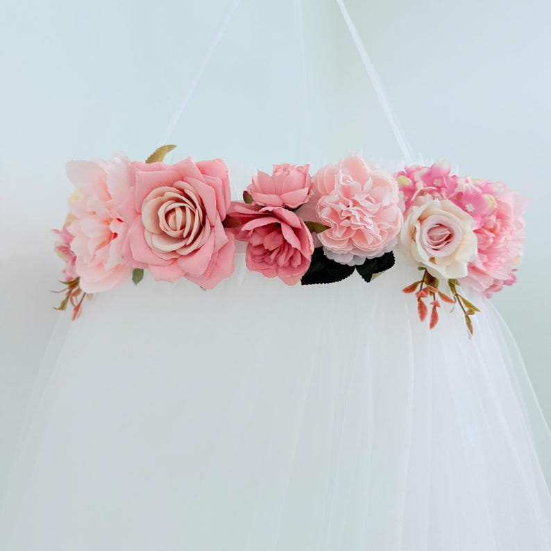 Puede incluir: Un dosel circular con una corona de flores artificiales en varios tonos de rosa, incluyendo rosa claro, rosa viejo y magenta. El arreglo floral est&aacute; sujeto a un cortinaje de tul blanco transparente que llega al suelo.