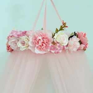 Puede incluir: Un dosel colgante con un marco circular adornado con un arreglo de flores artificiales en tonos rosa, crema y blanco. El dosel est&aacute; hecho de tela transparente de color rosa claro que cae hacia abajo desde el anillo cubierto de flores.
