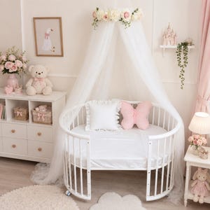 Puede incluir: Una cuna ovalada blanca con un dosel blanco transparente rematado con una corona de rosas rosas y blancas. Dentro de la cuna hay una almohada blanca cuadrada con volantes y una almohada rosa con forma de mariposa sobre un colch&oacute;n blanco.