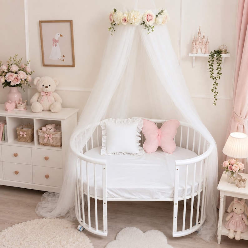 Puede incluir: Una cuna ovalada blanca con un dosel blanco transparente rematado con una corona de rosas rosas y blancas. Dentro de la cuna hay una almohada blanca cuadrada con volantes y una almohada rosa con forma de mariposa sobre un colch&oacute;n blanco.