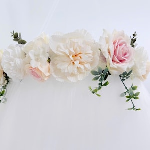 Puede incluir: Un adorno para dosel con un arreglo horizontal de flores artificiales, que incluye grandes peon&iacute;as color crema, rosas rosa suave y peque&ntilde;as flores blancas, acentuado con follaje verde sobre una base de tela blanca transparente.