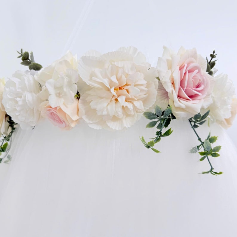 Puede incluir: Un adorno para dosel con un arreglo horizontal de flores artificiales, que incluye grandes peon&iacute;as color crema, rosas rosa suave y peque&ntilde;as flores blancas, acentuado con follaje verde sobre una base de tela blanca transparente.