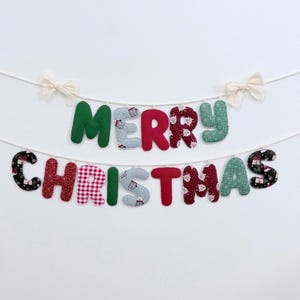 May include: A fabric banner spelling MERRY CHRISTMAS in two rows, suspended by white string with cream bows at the ends. The letters feature various patterns including solid green, red, plaid, and festive prints with small illustrations.
