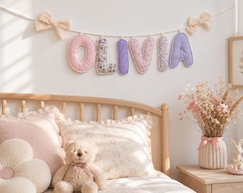 May include: A fabric letter banner spelling the name OLIVIA in pastel colors and floral patterns, suspended by a white cord with beige bows at each end. Each letter is individually padded and features distinct fabric designs.