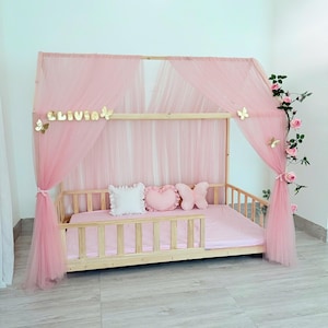 May include: A light wood-framed bed with a pink canopy and matching bedding. The canopy is draped with pink tulle and adorned with gold butterflies and the name "OLIVIA". Decorative pillows and floral accents add to the whimsical design.