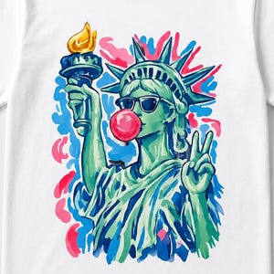 May include: White t-shirt featuring a colorful illustration of the Statue of Liberty wearing sunglasses and blowing a bubblegum bubble. The statue is holding a torch and making a peace sign with her fingers. The design includes blue, green, red, and pink accents.