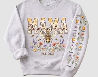 Puede incluir: Una sudadera gris claro con las palabras "MAMA Queenbee" en un diseño floral y de panal, con una ilustración de abeja. La manga tiene detalles florales y de nombres. También está presente el texto "EST. 2026".