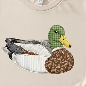 May include: A beige t-shirt featuring a detailed appliqué of a duck. The duck has a green checkered head, a yellow beak, and a brown floral patterned chest. The wings and body are made of various fabrics in shades of gray, white, and brown.
