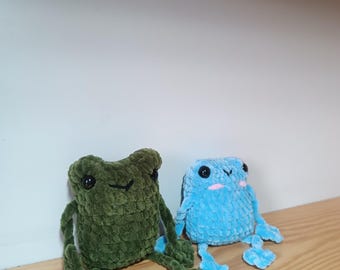 Op de afbeelding: Twee gehaakte kikker knuffels, een groene en een blauwe, zittend op een houten oppervlak.