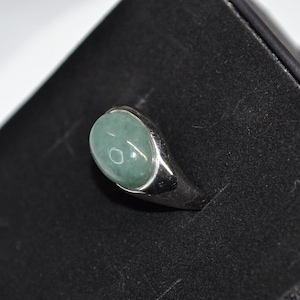 May include: A silver ring featuring a large, oval, pale green gemstone. The ring has a simple, polished silver band. The ring is presented on a black surface.