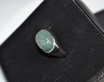 May include: A silver ring with a large, oval, pale green stone. The ring is set in a simple, polished silver band. The ring is displayed on a black surface.
