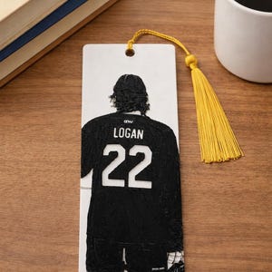 Puede incluir: Un marcap&aacute;ginas rectangular blanco con un gr&aacute;fico negro en relieve de un jugador de hockey visto desde atr&aacute;s. El jugador lleva una camiseta con el nombre LOGAN y el n&uacute;mero 22 en blanco. Una borla amarilla est&aacute; sujeta al agujero superior.
