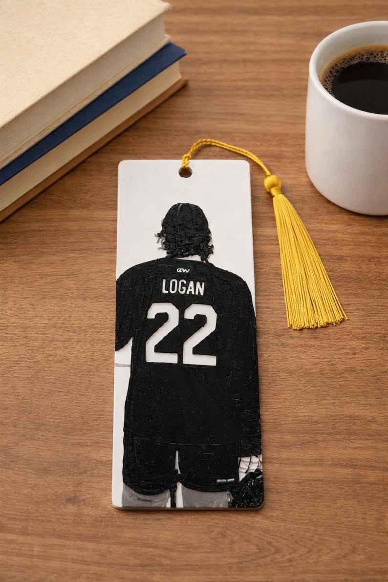 Puede incluir: Un marcap&aacute;ginas rectangular blanco con un gr&aacute;fico negro en relieve de un jugador de hockey visto desde atr&aacute;s. El jugador lleva una camiseta con el nombre LOGAN y el n&uacute;mero 22 en blanco. Una borla amarilla est&aacute; sujeta al agujero superior.