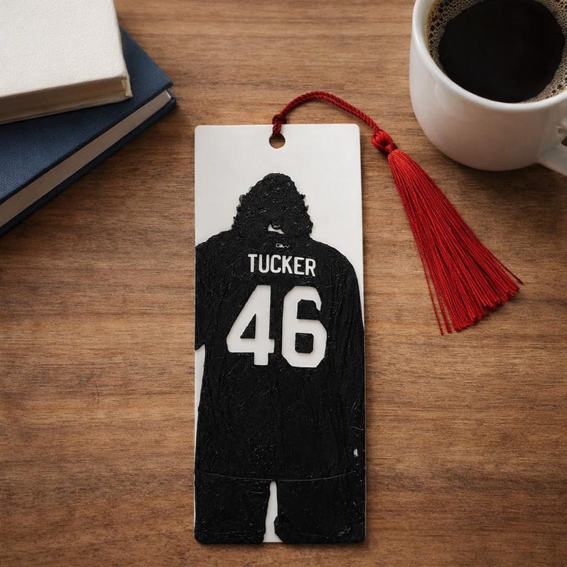 Puede incluir: Un marcap&aacute;ginas rectangular blanco con la silueta negra de una camiseta deportiva con el nombre TUCKER y el n&uacute;mero 46 impresos en blanco. El marcap&aacute;ginas incluye un agujero en la parte superior con una borla roja de hilos finos.