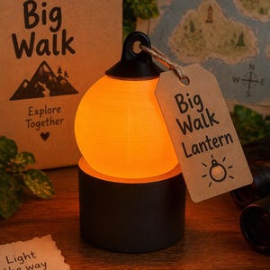 May include: A spherical orange glowing lantern sits on a black cylindrical base. A brown paper tag attached to the top loop reads Big Walk Lantern with a small icon. The lantern is accompanied by a cardboard box labelled Big Walk Explore Together.