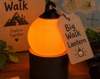 May include: A spherical orange glowing lantern sits on a black cylindrical base. A brown paper tag attached to the top loop reads Big Walk Lantern with a small icon. The lantern is accompanied by a cardboard box labelled Big Walk Explore Together.