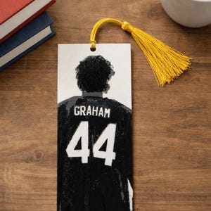Puede incluir: Un marcap&aacute;ginas rectangular con una ilustraci&oacute;n en blanco y negro de una persona vista desde atr&aacute;s, que lleva una camiseta negra con el nombre GRAHAM y el n&uacute;mero 44 impresos en blanco. Una borla amarilla brillante est&aacute; unida a la parte superior.