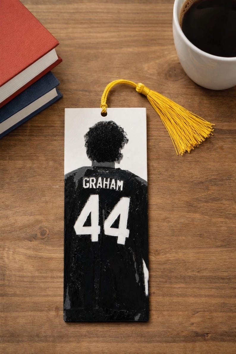 Puede incluir: Un marcap&aacute;ginas rectangular con una ilustraci&oacute;n en blanco y negro de una persona vista desde atr&aacute;s, que lleva una camiseta negra con el nombre GRAHAM y el n&uacute;mero 44 impresos en blanco. Una borla amarilla brillante est&aacute; unida a la parte superior.