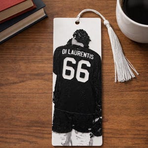 Puede incluir: Un marcap&aacute;ginas rectangular blanco con un relieve negro de una persona que lleva una camiseta con el nombre DI LAURENTIS y el n&uacute;mero 66 en la espalda. El marcap&aacute;ginas incluye un agujero en la parte superior con una borla blanca.