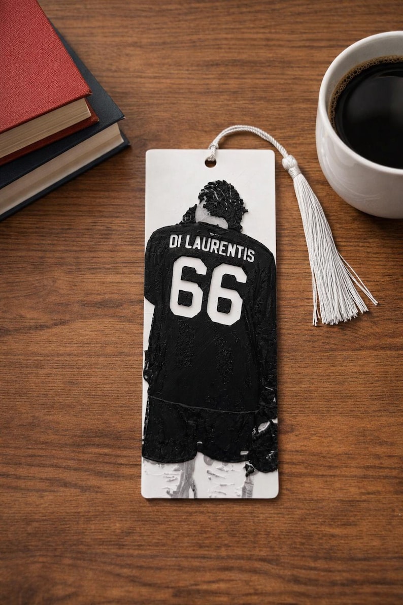 Puede incluir: Un marcap&aacute;ginas rectangular blanco con un relieve negro de una persona que lleva una camiseta con el nombre DI LAURENTIS y el n&uacute;mero 66 en la espalda. El marcap&aacute;ginas incluye un agujero en la parte superior con una borla blanca.