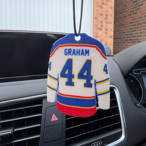 May include: A white, blue and red hanging car air freshener shaped like a hockey jersey. The jersey features the name GRAHAM in blue text above the number 44 in large blue digits. The sleeves and hem have blue and yellow stripes.