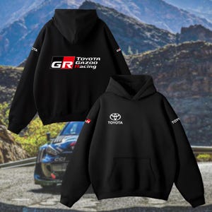 May include: Black hoodie featuring the Toyota Gazoo Racing logo and the Toyota logo on the sleeve. The background shows a mountain range and a race car.