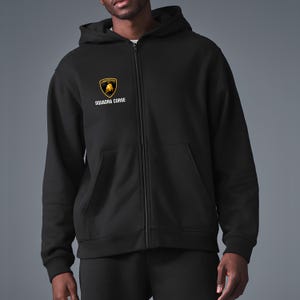 Può includere: Felpa nera con zip, cappuccio e tasche anteriori. La felpa presenta un logo Lamborghini oro e nero e le parole "SQUADRA CORSE" stampate sotto. La felpa è realizzata in materiale morbido.