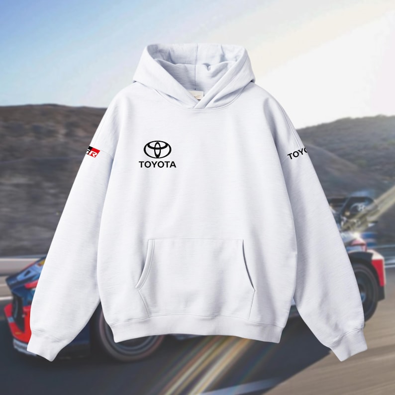 May include: White hoodie with a kangaroo pocket and hood. The front displays the Toyota logo in black, with "TOYOTA" beneath. The left sleeve has a "GR" logo. The background shows a race car on a track.