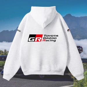 May include: A white hoodie with the Toyota Gazoo Racing logo in black, red, and white. The sweatshirt has a drawstring hood and long sleeves. The logo is on the back. The word "OTA" is printed on the left sleeve.