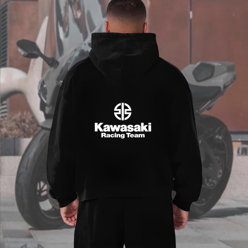 May include: Black hoodie with the Kawasaki Racing Team logo in white on the back. The logo features a stylized emblem above the text. The person is wearing black pants. A motorcycle is visible in the background.