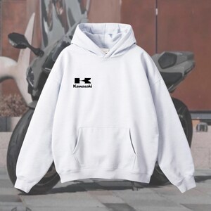 May include: White hoodie with a kangaroo pocket and a hood. The black Kawasaki logo is on the chest. The background features a motorcycle.