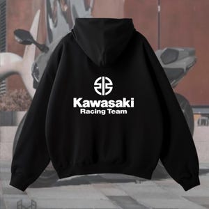 May include: Black hoodie with the Kawasaki Racing Team logo in white. The logo features a stylized emblem above the text. The hoodie has a hood and long sleeves. The background shows a motorcycle.