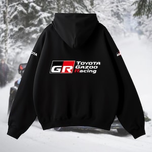 May include: Black hooded sweatshirt with the "TOYOTA GAZOO Racing" logo in white, red, and black. The "GR" logo is on the front. The sweatshirt has a hood and long sleeves. The background shows a snowy scene with a vehicle.