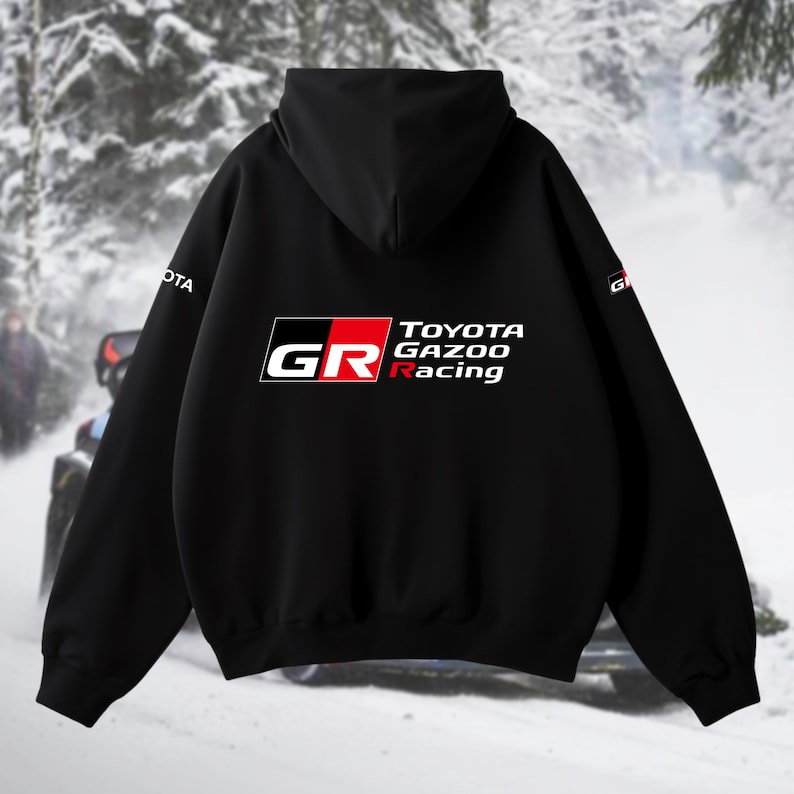 May include: Black hooded sweatshirt with the "TOYOTA GAZOO Racing" logo in white, red, and black. The "GR" logo is on the front. The sweatshirt has a hood and long sleeves. The background shows a snowy scene with a vehicle.