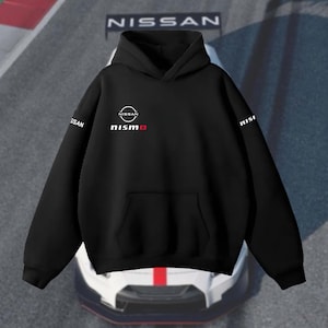 Op de afbeelding: Zwarte hoodie met een voorzak en een capuchon. Het Nissan-logo en het woord "nismo" in het rood staan op de voorkant. De mouwen hebben het woord "nissan" in het wit gedrukt. De achtergrond toont een auto.