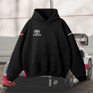 May include: Black hoodie with the Toyota logo in white on the chest. The word "TOYOTA" is also printed in white. The left sleeve has a red and white "GR" logo. The hoodie has a front pocket and a hood.