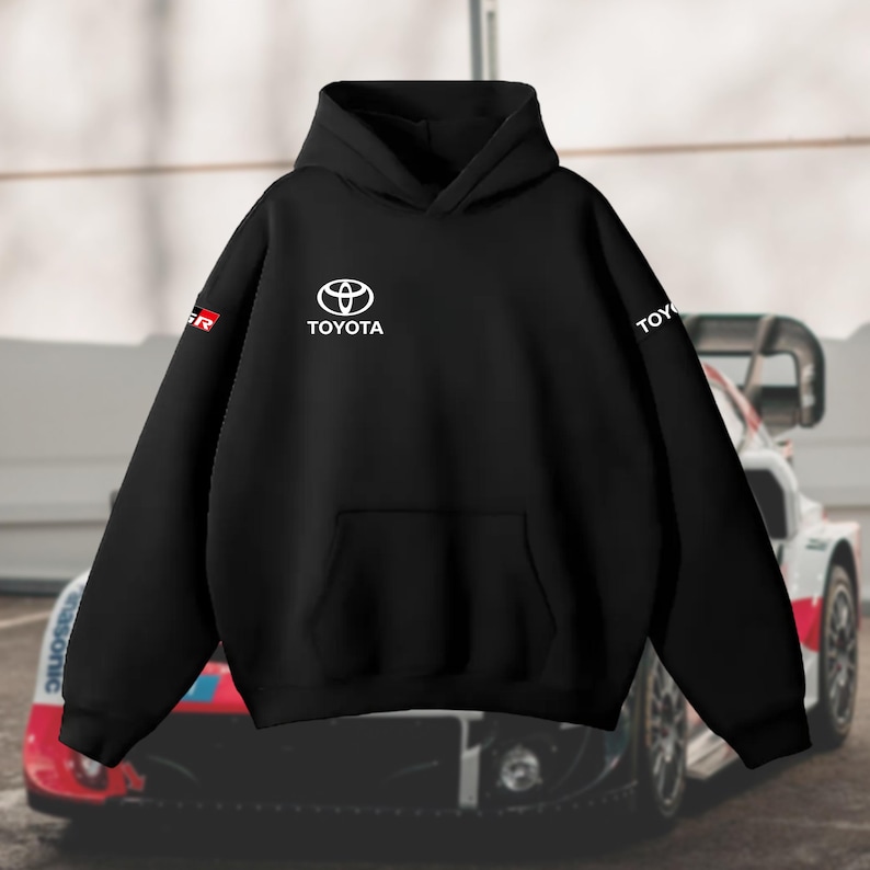 May include: Black hoodie featuring the Toyota logo in white on the chest, with the word "TOYOTA" also in white. The left sleeve displays a red and white "GR" logo. The hoodie includes a front pocket and a hood.