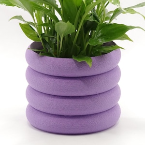 Può includere: Una rigogliosa pianta verde con foglie grandi è piantata in un vaso impilato, rotondo, color lavanda. Il vaso ha una superficie strutturata e un design moderno. Le foglie della pianta sono lucide, creando un contrasto sorprendente.