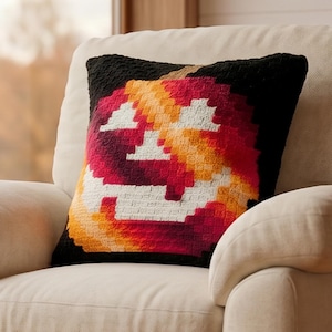 May include: A square throw pillow featuring a pixelated jack-o'-lantern design. The pumpkin is rendered in shades of orange, red, and yellow against a solid black background, displaying a classic carved face with triangular eyes and a wide grin.