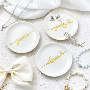 May include: Three white ceramic ring dishes with gold script names: "Jenna", "Sindy", and "Olivia". The "Sindy" dish has pearl earrings and a "Bride" hair clip. Jewellery and a white bow are also visible.