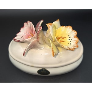 May include: A white ceramic trinket box with a lid, adorned with sculpted, colorful flowers. The flowers are pink, yellow, and orange, with detailed petals. The box has a small, oval shape and a glossy finish.