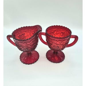 May include: A red glass creamer and sugar bowl set. The set features a detailed grape design and scalloped edges. Both pieces have handles and sit on a pedestal base. The creamer has a spout for pouring.