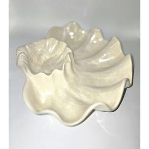 May include: Off-white, clam-shaped serving dish. The dish has a large, wavy outer shell and a smaller, bowl-shaped inner section. The surface has a glossy finish, and the overall design is reminiscent of the ocean.