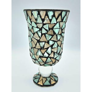 May include: A decorative glass vase with a mosaic design. The vase is adorned with triangular pieces in shades of beige, teal, and clear glass, set against a black background. The vase has a round base and a clear glass ball supporting the main body.