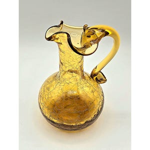 May include: A vintage amber-colored glass pitcher with a crackle finish. The pitcher has a rounded base, a slender neck, and a ruffled rim. It features a curved handle and is likely used for serving beverages.
