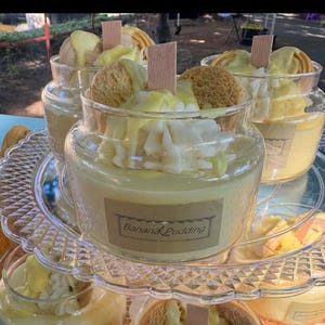 May include: Glass jars filled with banana pudding, topped with whipped cream, cookies, and wooden sticks. The jars are arranged on a tiered, clear serving tray. The label on the jar reads "Banana Pudding".